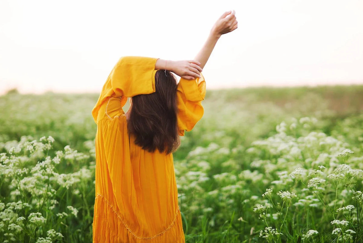 Woman in a golden-yellow dress standing in a wildflower field at golden hour, back to camera, one arm raised through her hair as if letting go of something heavy. A quiet moment of lightness returning.