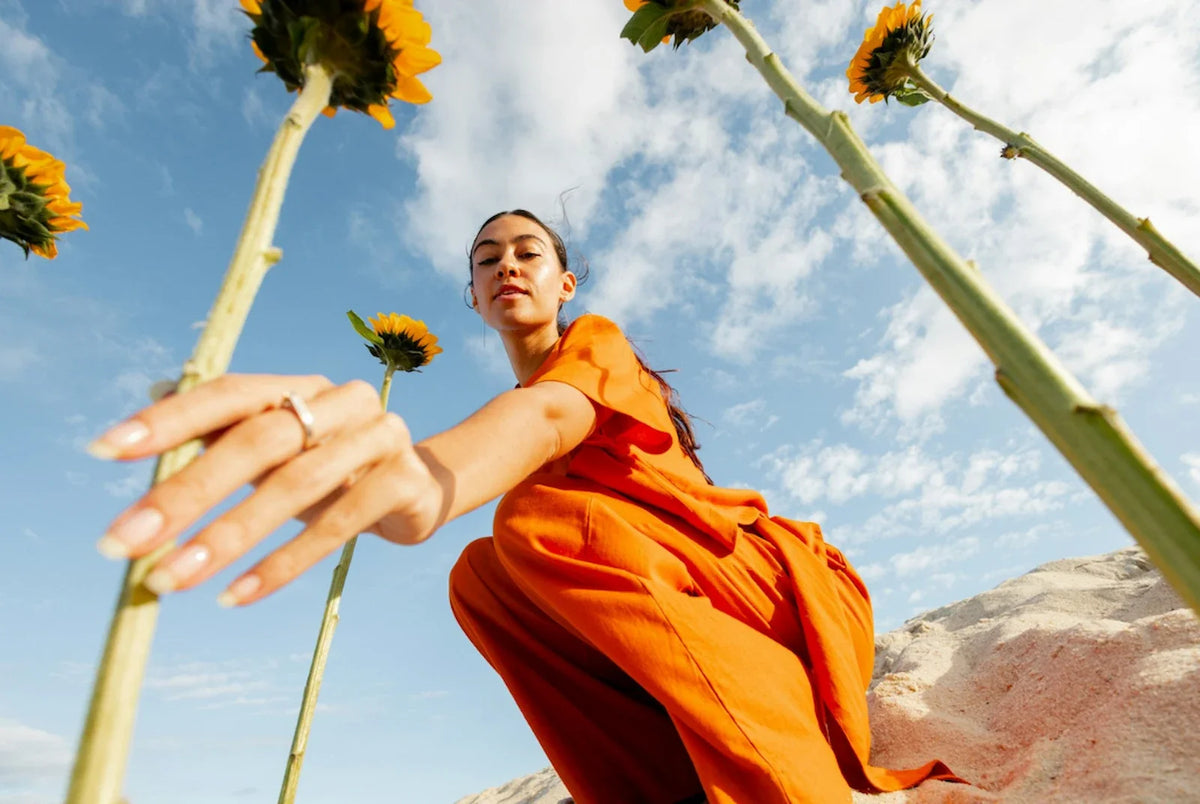 Woman in an orange dress crouching low among tall sunflowers, reaching her hand directly toward the camera, sky and clouds behind her.
