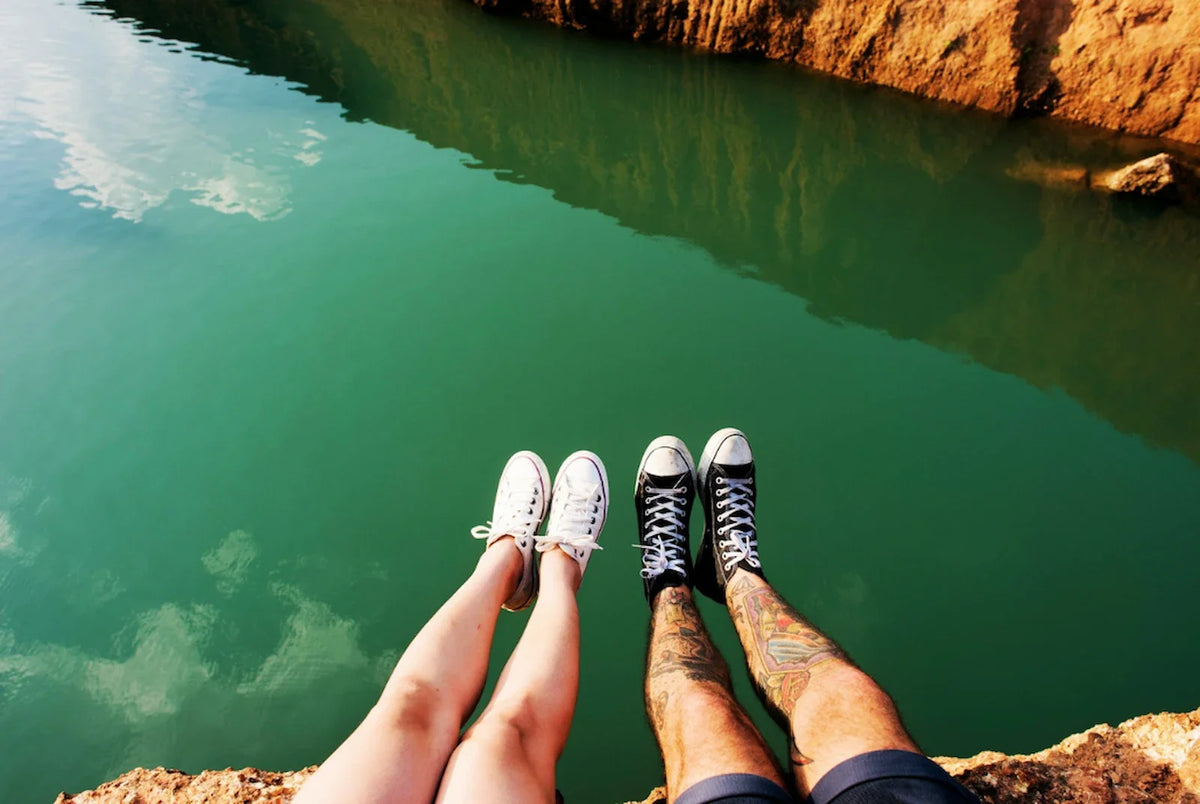 Two people sitting side by side on a rocky ledge, legs dangling over turquoise water below, sneakers visible on both.