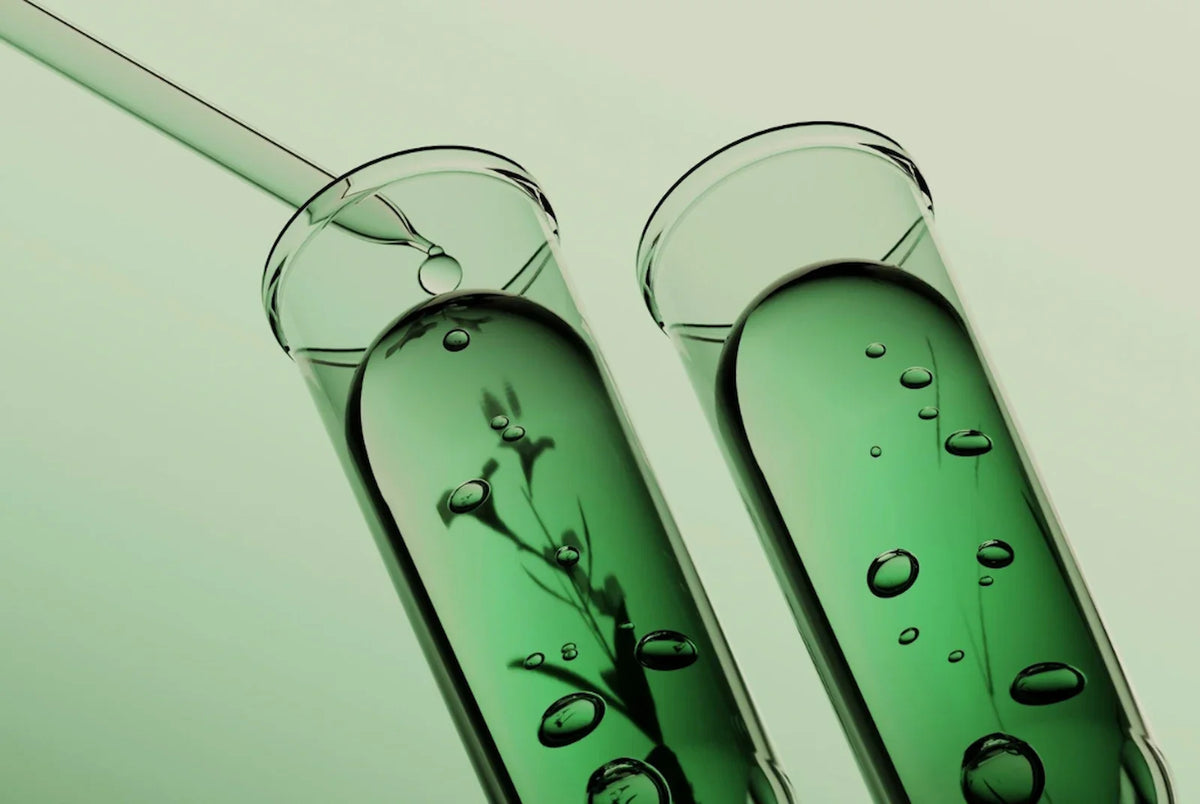 Two test tubes filled with green liquid side by side, a pipette adding a drop into the left one, faint plant-like shadows visible inside it while the other tube appears clear.