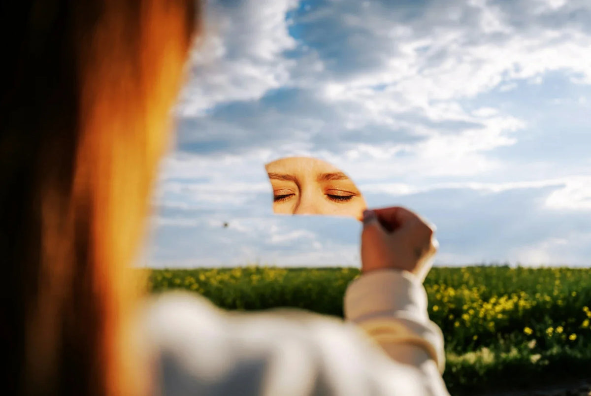 Woman from behind holding up a small round mirror against the sky, her closed eyes reflected in it, a blurred field of yellow flowers behind her.