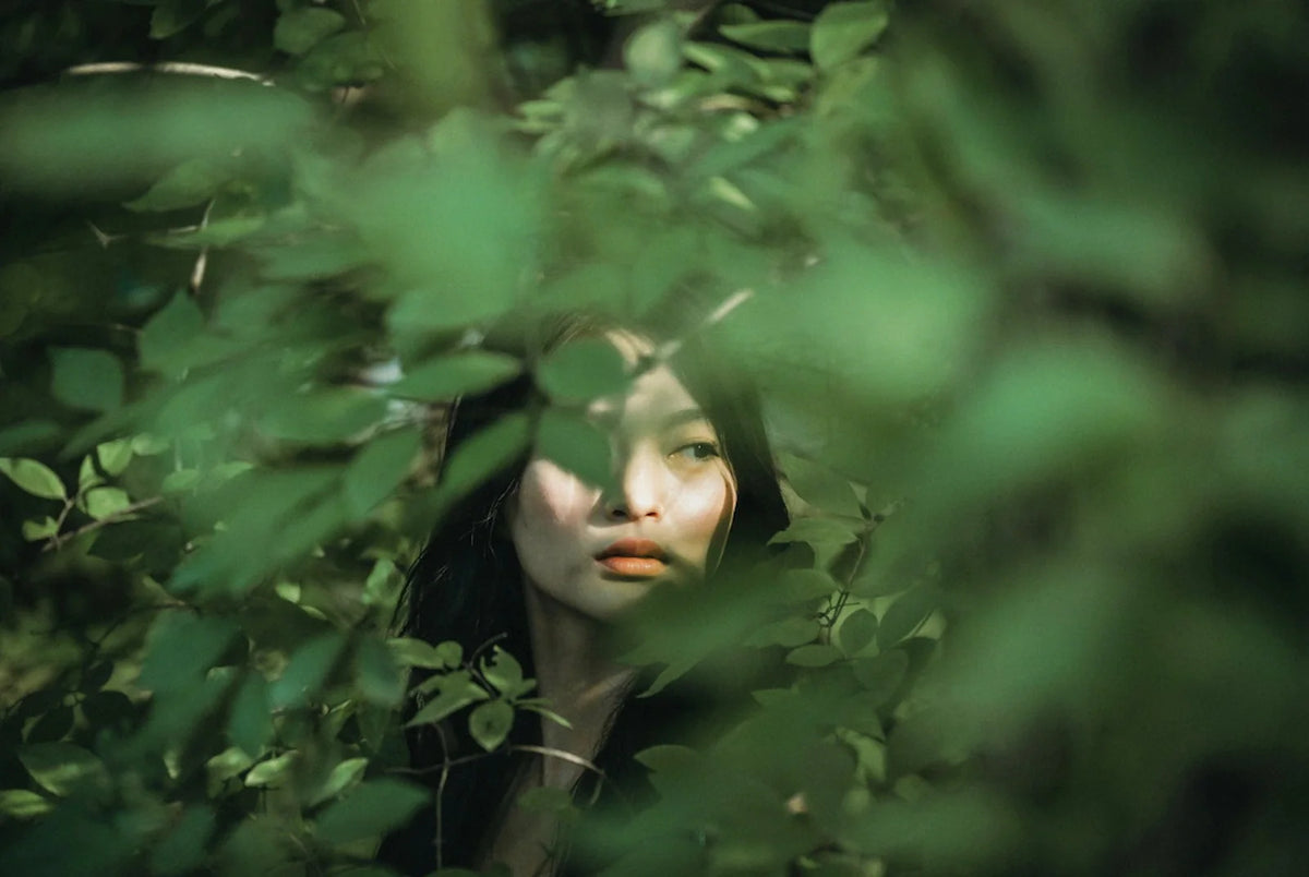 Woman's face half-hidden behind dense green leaves, eyes lowered, as if pulling back from being seen.