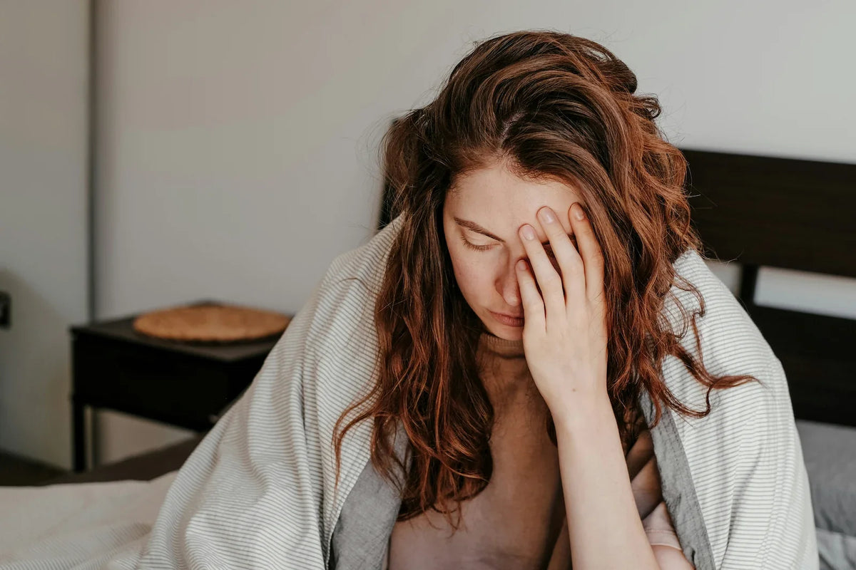 Woman wrapped in a blanket on her bed with a hand pressed to her forehead, representing a PMDD spiral