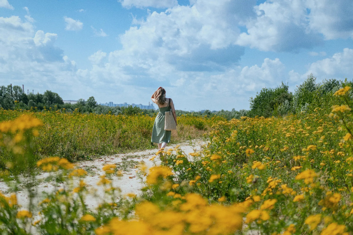 A woman in a long green dress stands on a sandy path in a flower field under a blue sky with clouds, surrounded by yellow wildflowers.